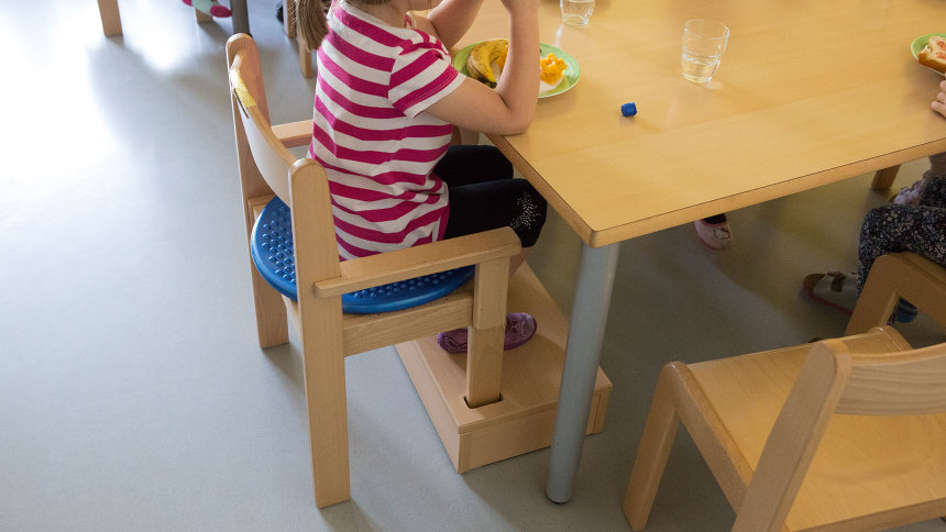 Ein Mädchen sitzt auf einem Stuhl mit Armlehnen am Tisch.