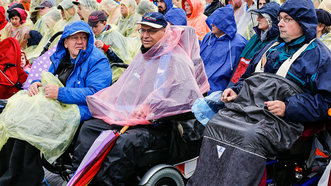 Publikum bei einer Freiluftveranstaltung; im Vordergrund mehrere Besucher im Rollstuhl. Alle Menschen tragen Regenkleidung.