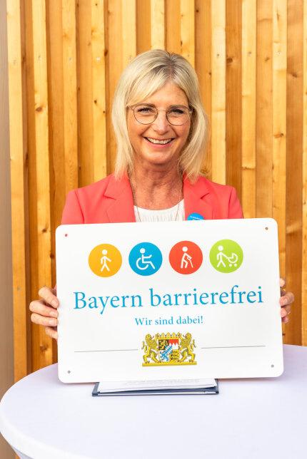 Vor einer Holzwand zeigt Sozialministerin Ulrike Scharf das Signet „Bayern barrierefrei – Wir sind dabei!“.