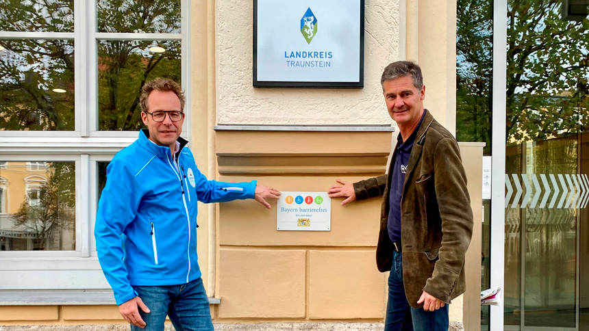 Zwei Personen zeigen auf ein an einer Hauswand angebrachtes Signet „Bayern barrierefrei“.