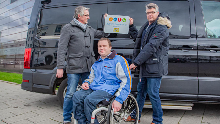 Gruppenbild: Übergabe des Signets „Bayern barrierefrei“ vor einem Kleinbus.