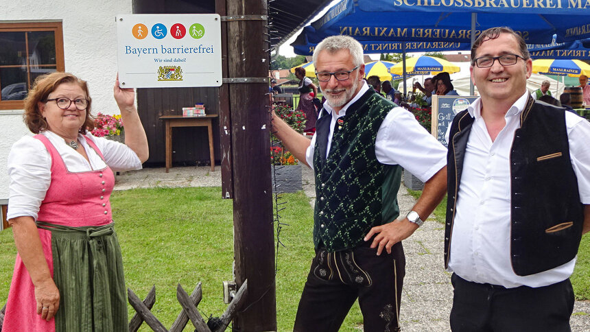 Drei Personen in Tracht mit dem Signet „Bayern barrierefrei“. 