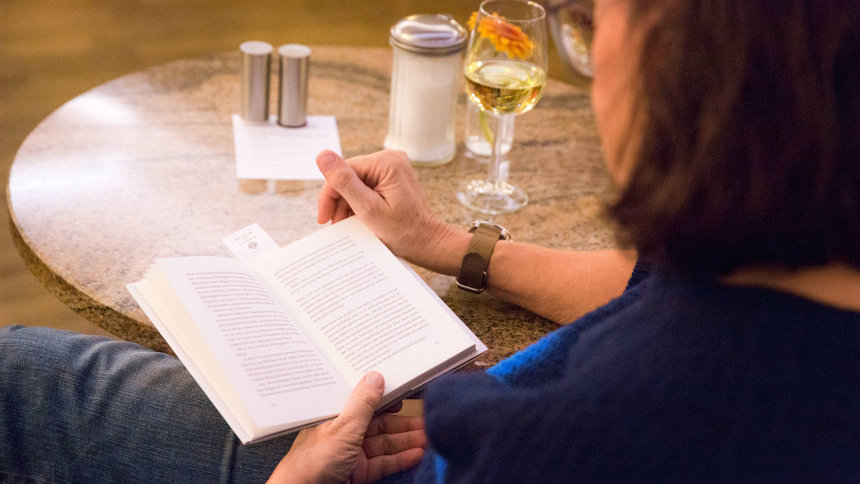 Eine Frau sitzt mit Buch und Weinglas an einem Bistrotisch.