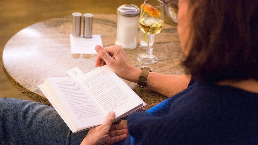 Eine Frau sitzt mit Buch und Weinglas an einem Bistrotisch.