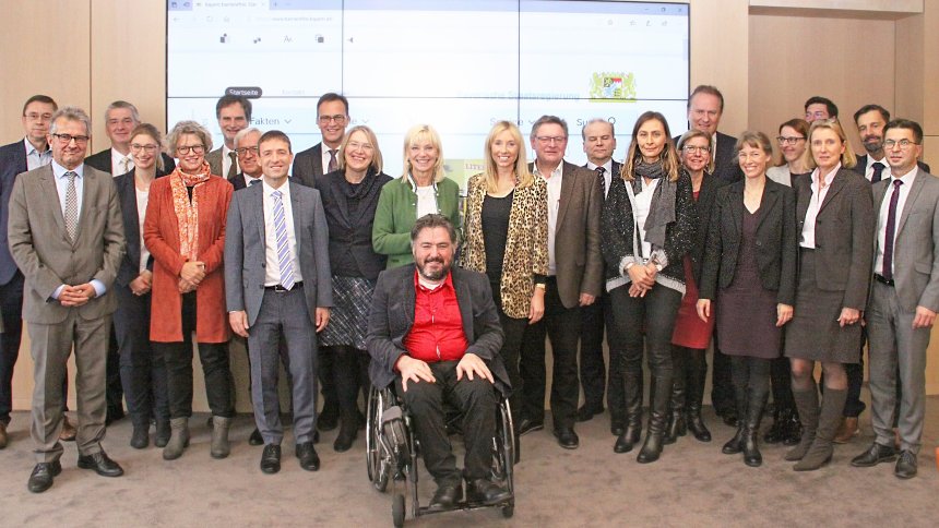Gruppenbild im Bayerischen Sozialministerium: Sitzungsteilnehmende stehend, vorne ein Teilnehmer im Rollstuhl.
