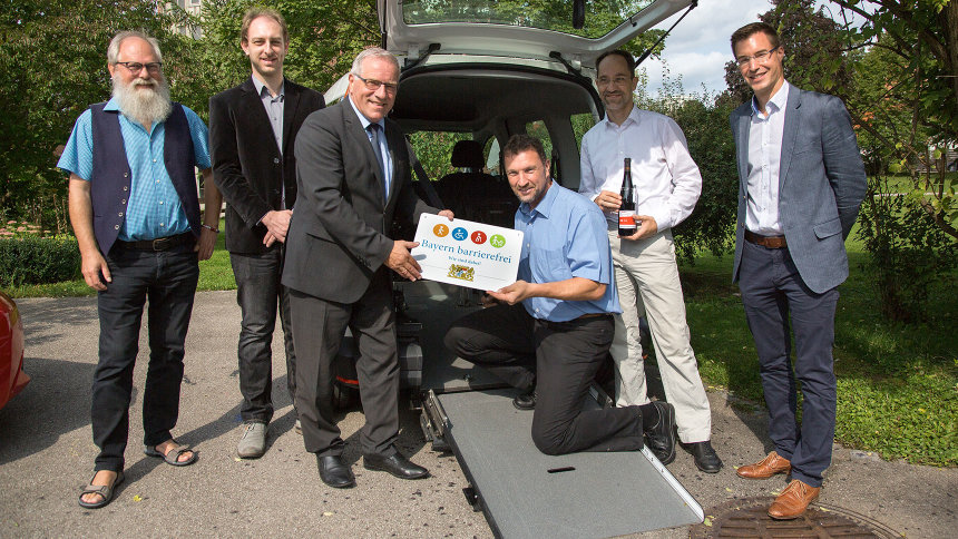 Gruppenbild: Übergabe des Signets „Bayern barrierefrei“ vor der Rampe eines umgebauten Autos.