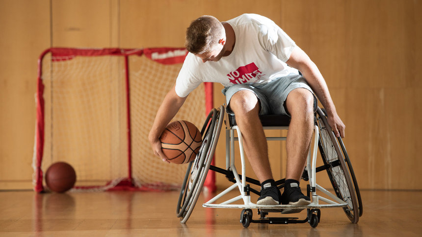 In einer Sporthalle: Ein junger Mann im Sportrollstuhl hebt einen Basketball auf.