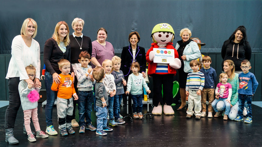 Gruppenbild: Erwachsene, Kinder und lebensgroßes Playmobil-Männchen bei Übergabe des Signets „Bayern barrierefrei“.