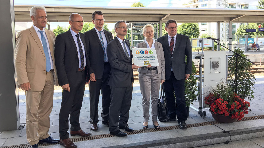 Gruppenbild: Übergabe des Signets „Bayern barrierefrei“ an einem Bahnsteig.