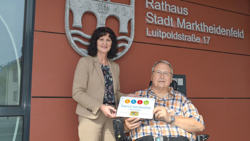 Zwei Personen mit dem Signet „Bayern barrierefrei“ vor dem Rathaus von Marktheidenfeld.