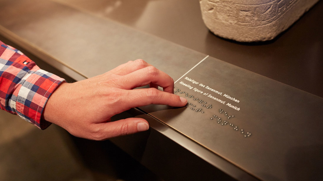 Museumsbesucherin vor einem Ausstellungsstück. Sie tastet die Erläuterung zum Austellungstück in Braille-Schrift ab.