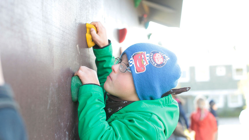 Nahaufnahme: Ein Bub mit Brille hangelt sich konzentriert an Griffen einer Kletterwand empor. 