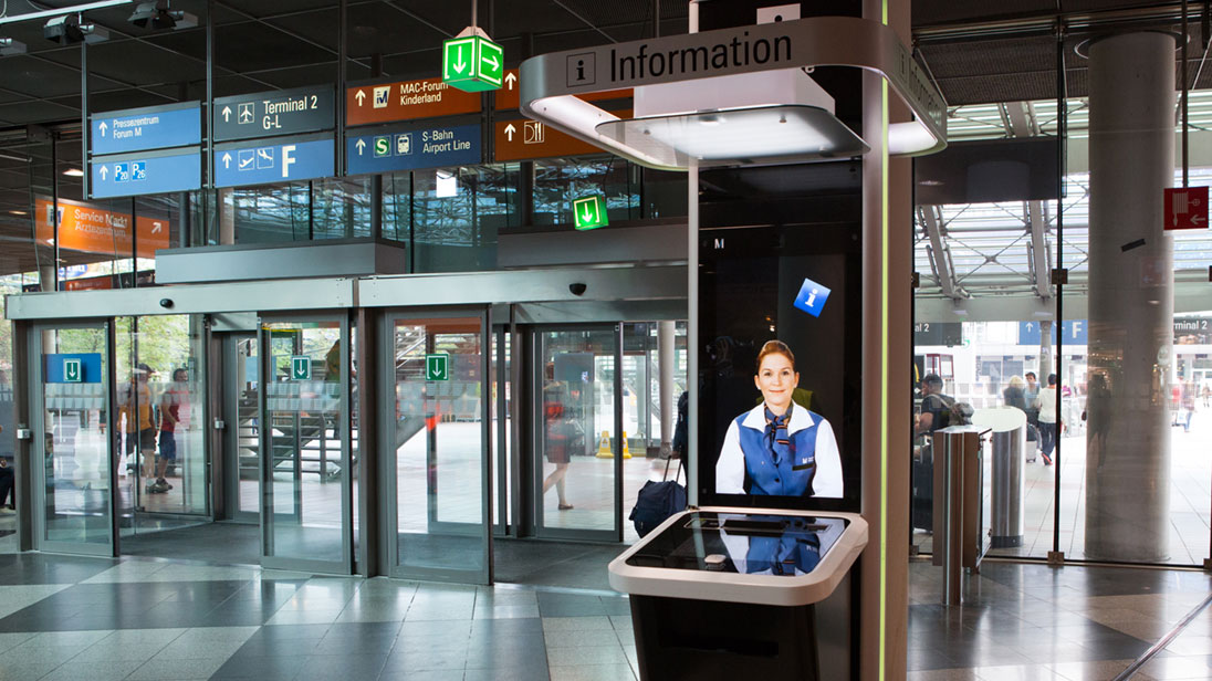 Blick in ein Flughafengebäude; im Vordergrund ein interaktives Info-Terminal.