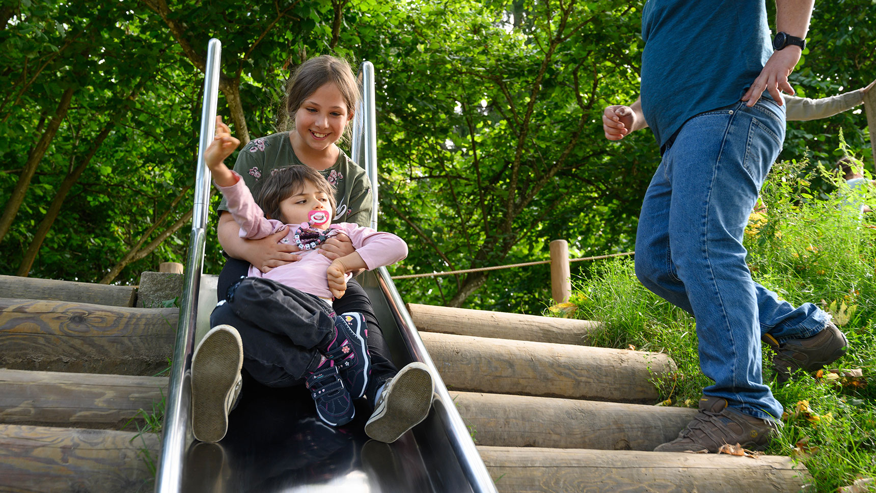 Lea und Hanna sitzen auf einer Rutsche. Ihr Vater steht daneben auf einer Treppe aus Holzstämmen.