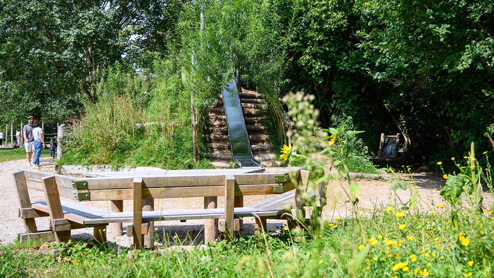 Blick aufs Spielplatzgelände: vorne eine Wiese mit blühenden Blumen, dahinter ein Picknicktisch.