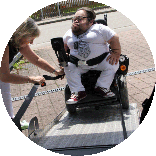 Benedikt Lika steht mit seinem Rollstuhl auf der Rampe eines Autos. Eine Frau befestigt das Zugseil.
