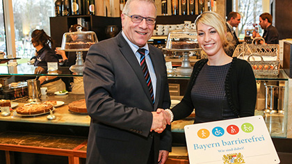 Sozialstaatssekretär Johannes Hintersberger übergibt das Signet „Bayern barrierefrei“ an Melanie Muth vom Restaurant „Ella“. 