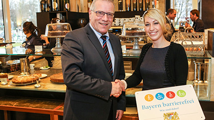 Sozialstaatssekretär Johannes Hintersberger übergibt das Signet „Bayern barrierefrei“ an Melanie Muth vom Restaurant „Ella“.