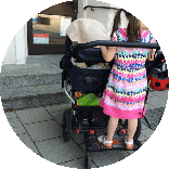 Ein Doppelkinderwagen vor zwei Stufen, die zu einer Ladentür führen. Auf dem Rollbrett des Wagens steht ein kleines Mädchen.