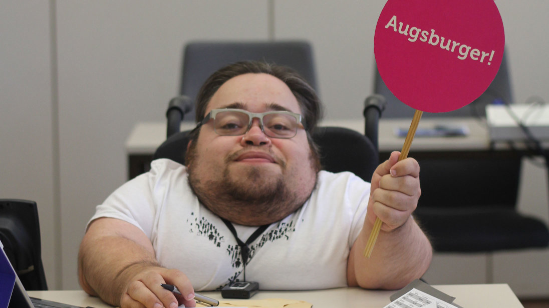 Benedikt Lika an einem Arbeitsplatz. Er trägt ein weißes T-Shirt und hält ein rundes, signalrotes Schild mit der Aufschrift „Augsburger“ hoch.