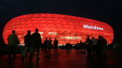 Besucher strömen zur Münchner Allianz Arena, die in der Dunkelheit rot leuchtet.