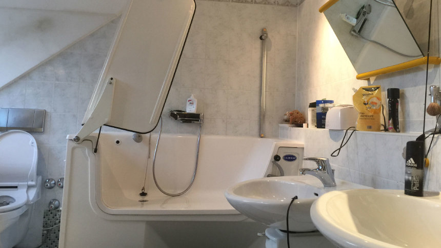 Badezimmer mit Dusch-WC, absenkbarem Waschbecken, kippbarem Spiegel und einer Badewanne mit Tür