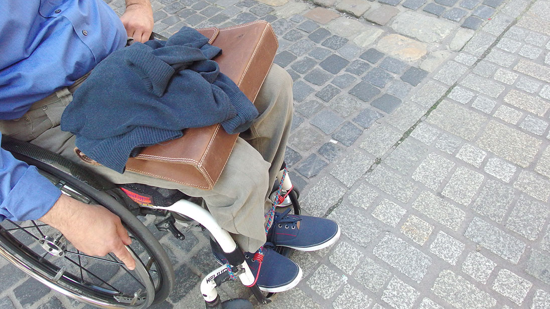 Ein Mann steht mit seinem Rollstuhl in einer Fußgängerzone, am Übergang von grober zu abgeflachter, rollstuhlfreundlicher Pflasterung. 