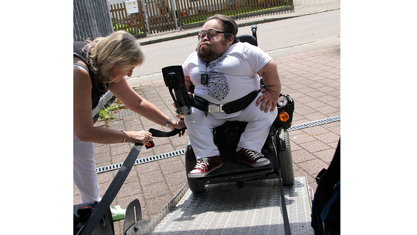 Benedikt Lika steht mit seinem Rollstuhl auf der Rampe eines Autos. Eine Frau befestigt das Zugseil.