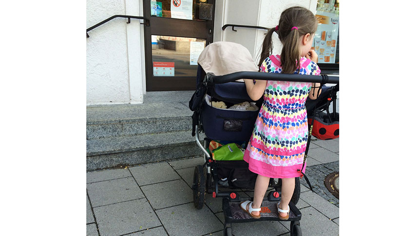 Ein Doppelkinderwagen vor zwei Stufen, die zu einer Ladentür führen. Auf dem Rollbrett des Wagens steht ein kleines Mädchen.