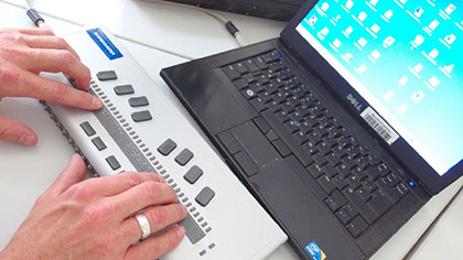 Notebook mit angeschlossenem Braille-Display. Die Hände des Benutzers liegen auf der Braille-Zeile.