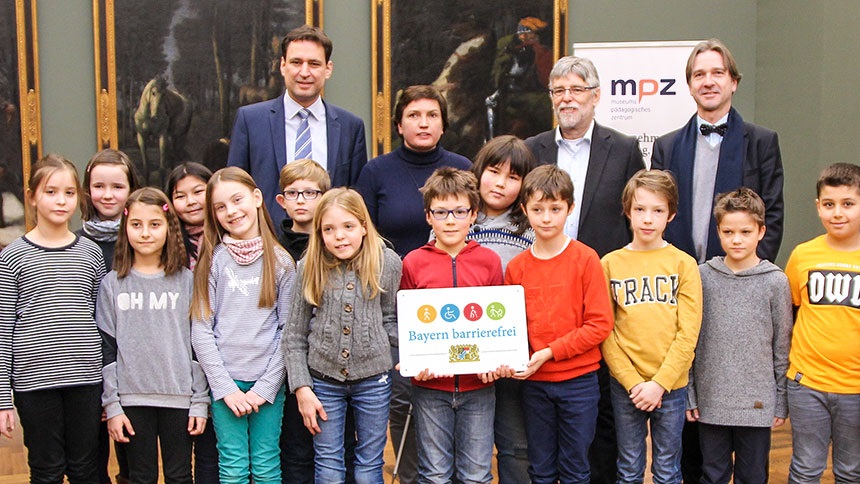 Gruppenbild: Grundschulkinder und vier Erwachsene präsentieren das Signet „Bayern barrierefrei“.