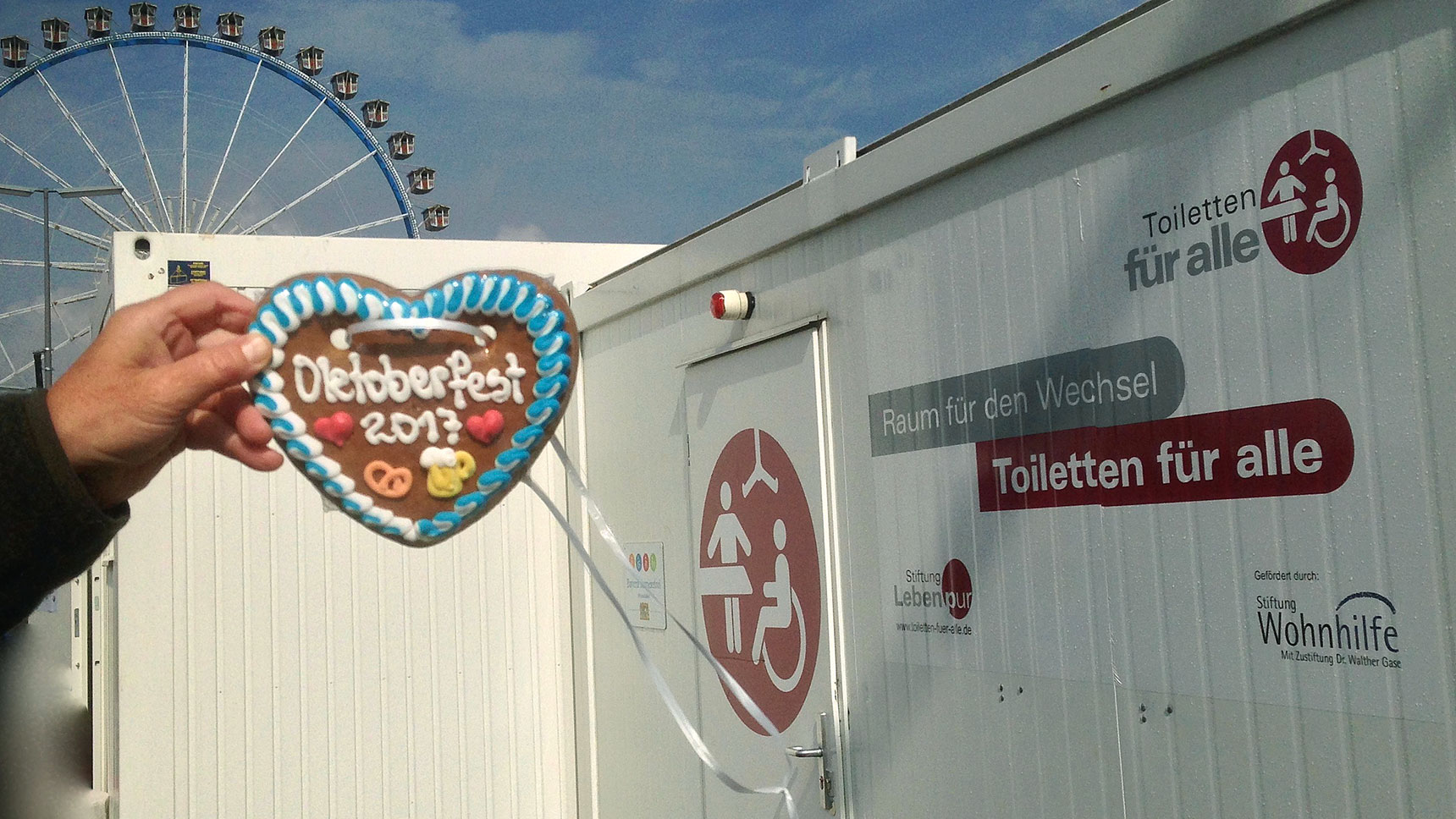 Eine Hand hält ein Lebkuchenherz mit der Aufschrift „Oktoberfest 2017“ in die Kamera, daneben der Toilettencontainer, dahinter das Riesenrad.