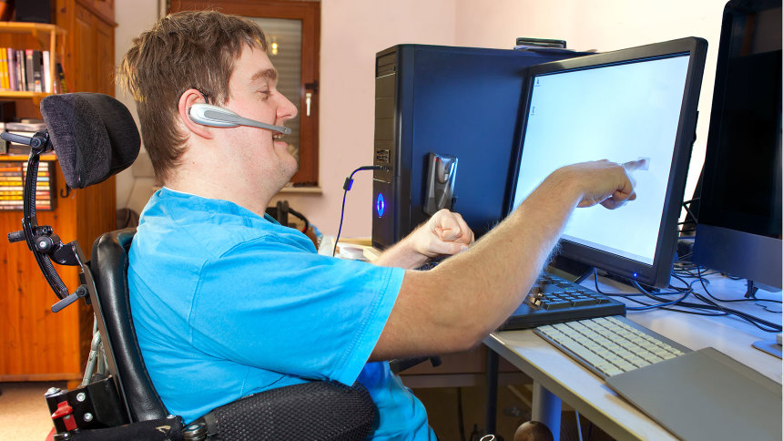 Ein junger Mann im Rollstuhl bedient einen Computer über ein Touch-Display und ein Headset. 