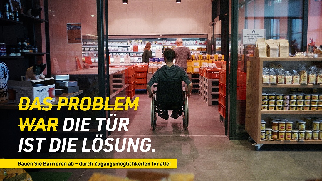 Ein Rollstuhlfahrer fährt durch eine Eingangstür in ein Geschäft. Auf dem Bild ist in Großbuchstaben die Aufschrift: 