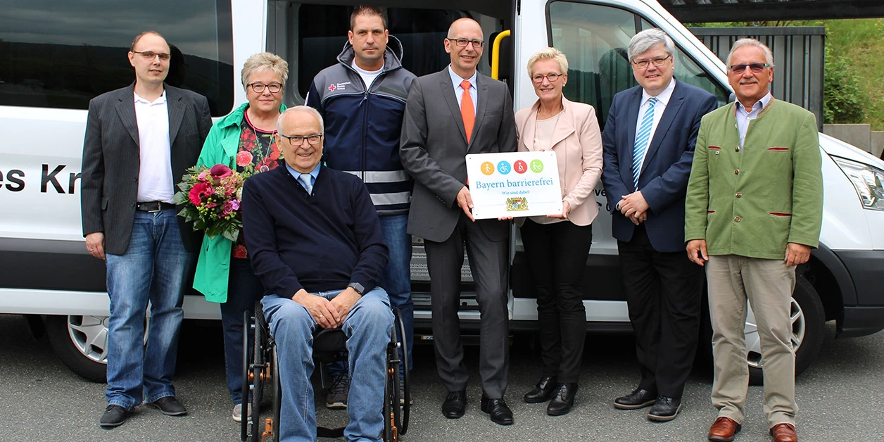 Gruppenbild: Übergabe des Signets „Bayern barrierefrei“ vor einem Kleinbus.