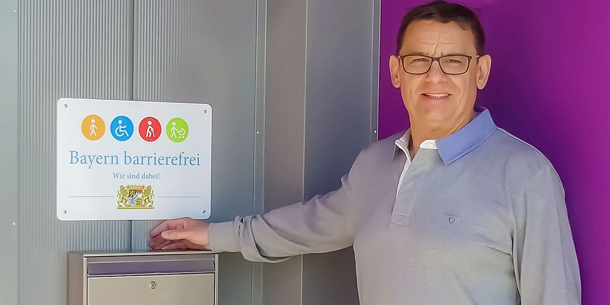 Geschäftsführer Bodo Schrödel steht neben dem im Eingangsbereich befestigten Signet „Bayern barrierefrei”.