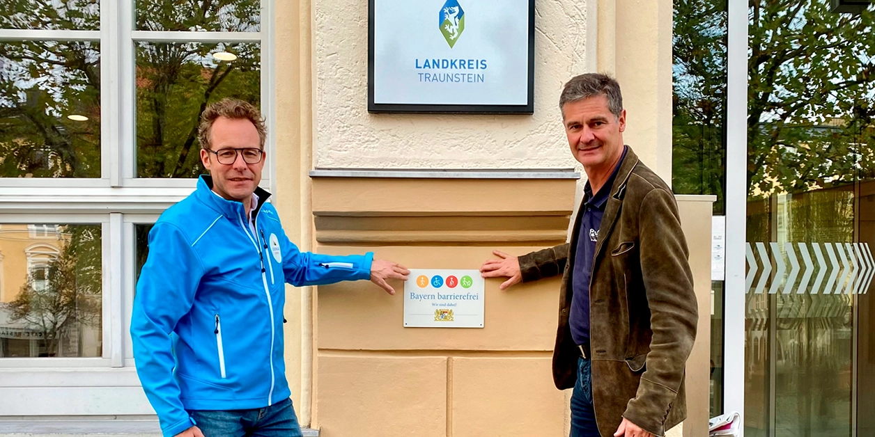 Zwei Personen zeigen auf ein an einer Hauswand angebrachtes Signet „Bayern barrierefrei”