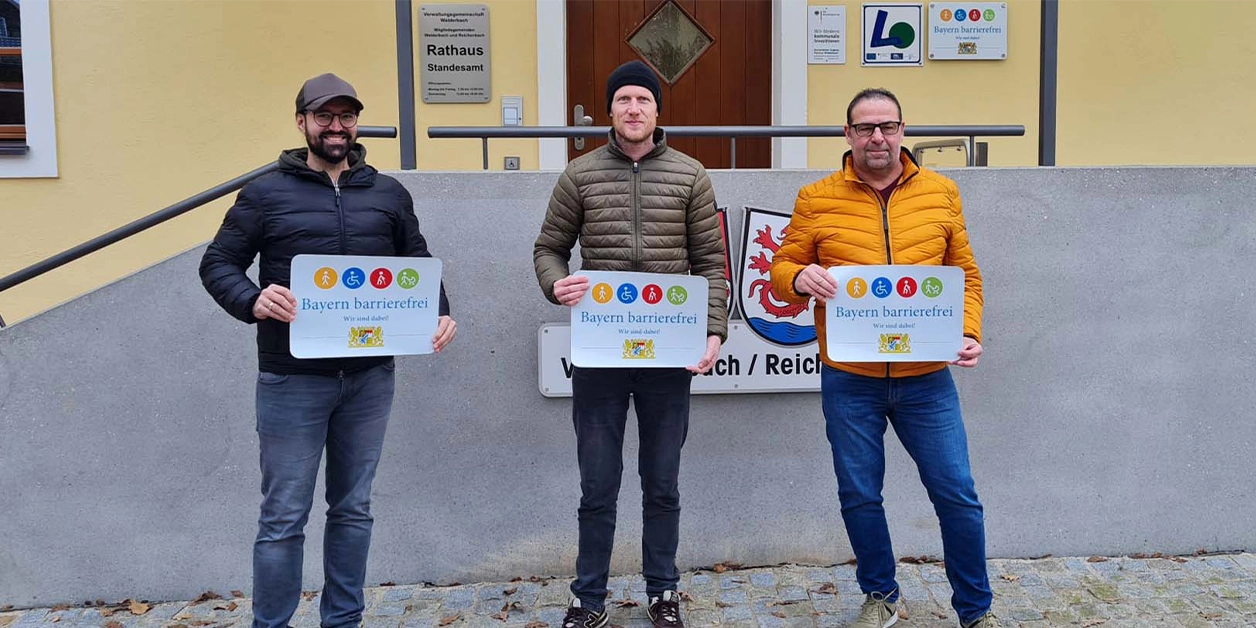 Vor dem Aufgang zu einem Gebäude: Drei Männer präsentieren das Signet „Bayern barrierefrei“.