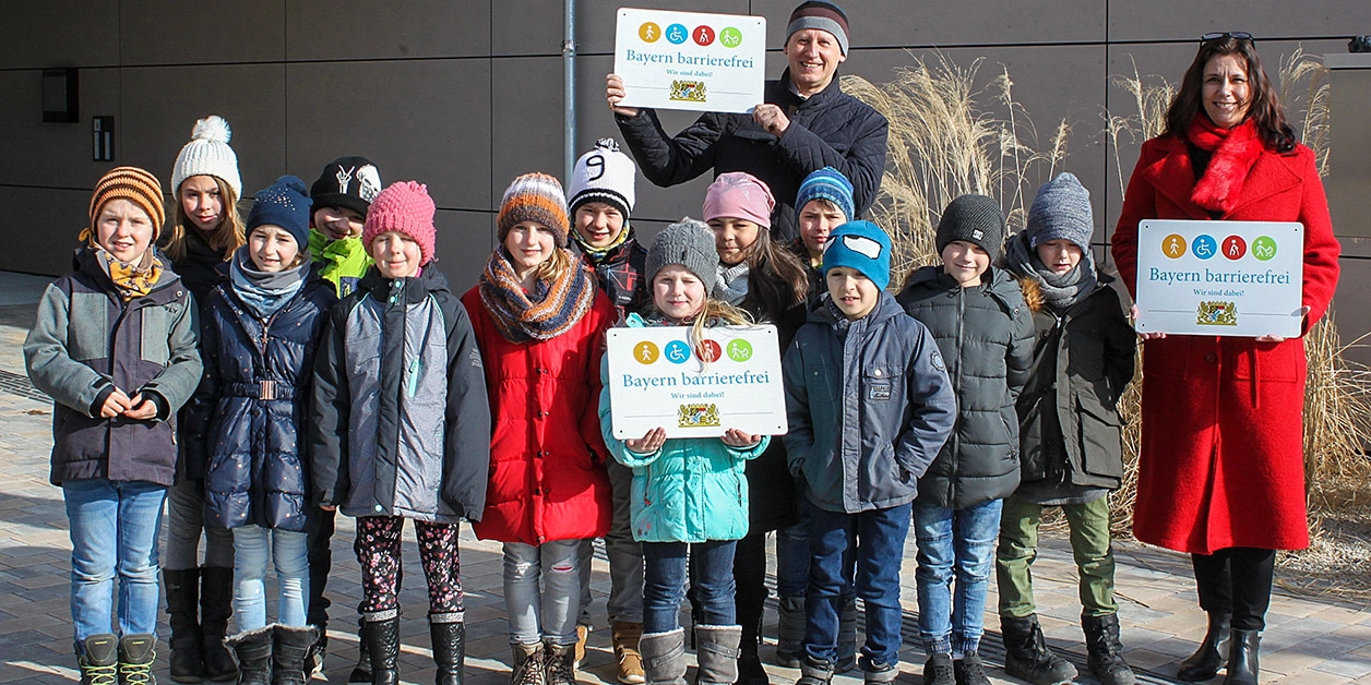 Gruppenbild: Kinder und Erwachsene zeigen Signets „Bayern barrierefrei”.