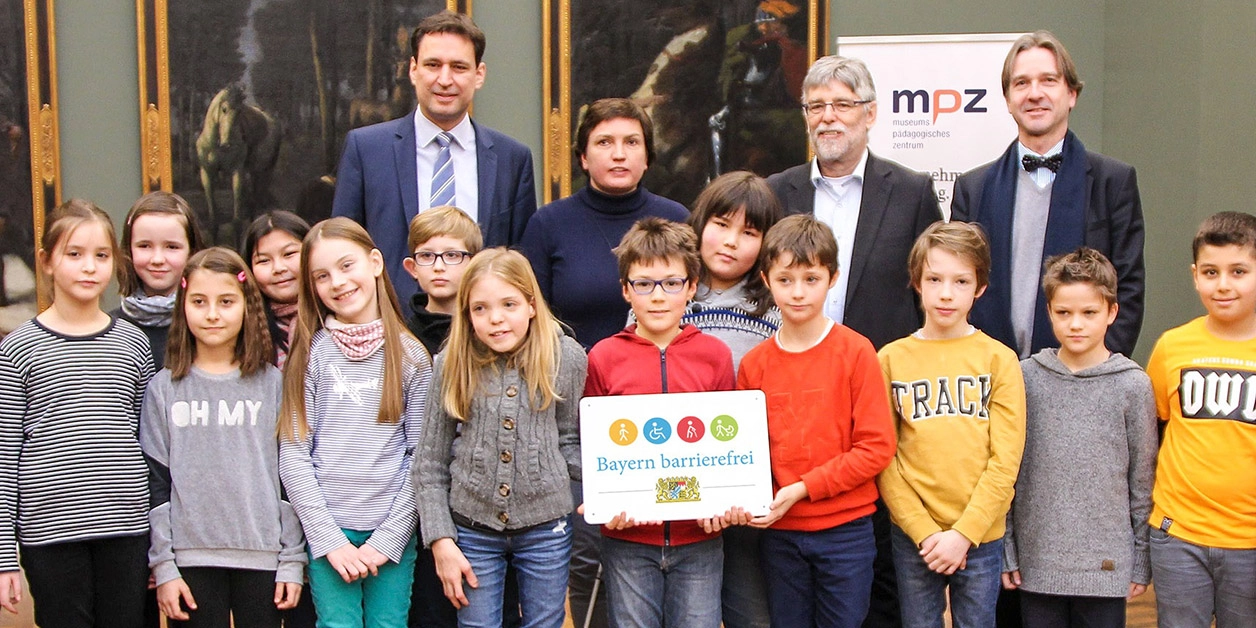 Gruppenbild: Grundschulkinder und vier Erwachsene präsentieren das Signet „Bayern barrierefrei”.