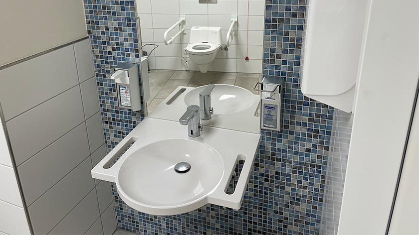 Blick in ein barrierefreies WC: vorne ein Waschtisch, dahinter spiegelt sich eine Toilette mit Haltegriffen und Notknopf. 