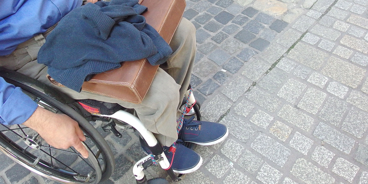 Ein Mann steht mit seinem Rollstuhl in einer Fußgängerzone, am Übergang von grober zu abgeflachter, rollstuhlfreundlicher Pflasterung. 