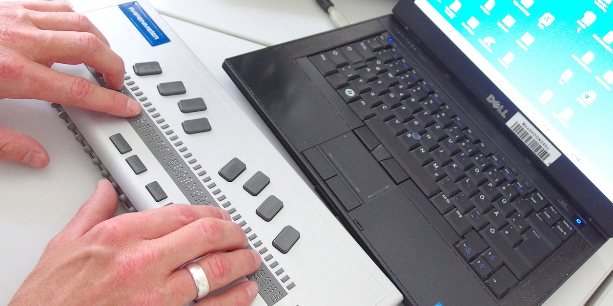 Ein Notebook, davor eine Braillezeile, auf der Finger beider Hände entlangtasten.