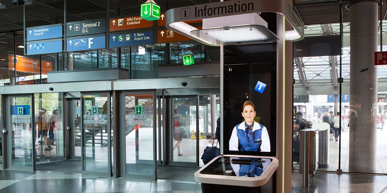 Blick in ein Flughafengebäude; im Vordergrund ein interaktives Info-Terminal.