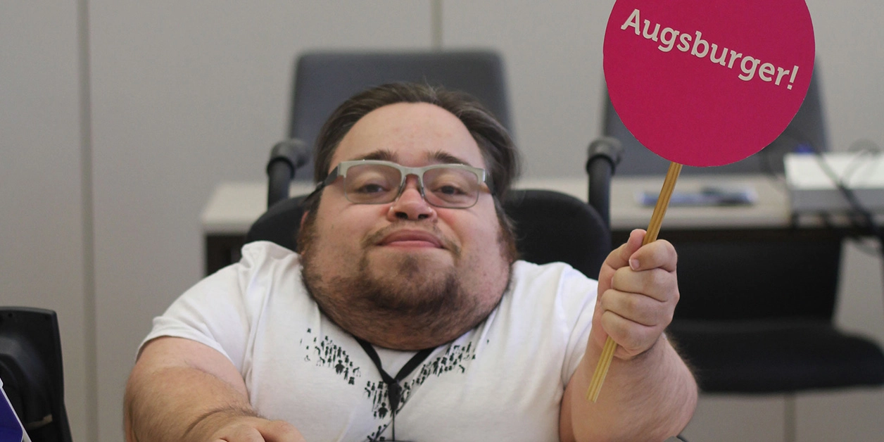 Benedikt Lika an einem Arbeitsplatz. Er trägt ein weißes T-Shirt und hält ein rundes, signalrotes Schild mit der Aufschrift „Augsburger“ hoch.
