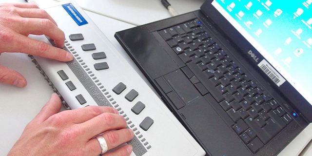 Ein Notebook, davor eine Braillezeile, auf der Finger beider Hände entlangtasten.