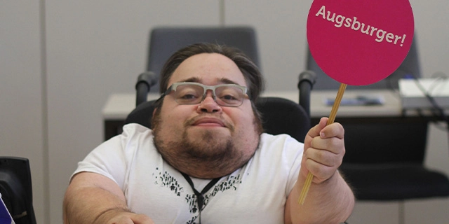 Benedikt Lika an einem Arbeitsplatz. Er trägt ein weißes T-Shirt und hält ein rundes, signalrotes Schild mit der Aufschrift „Augsburger“ hoch.