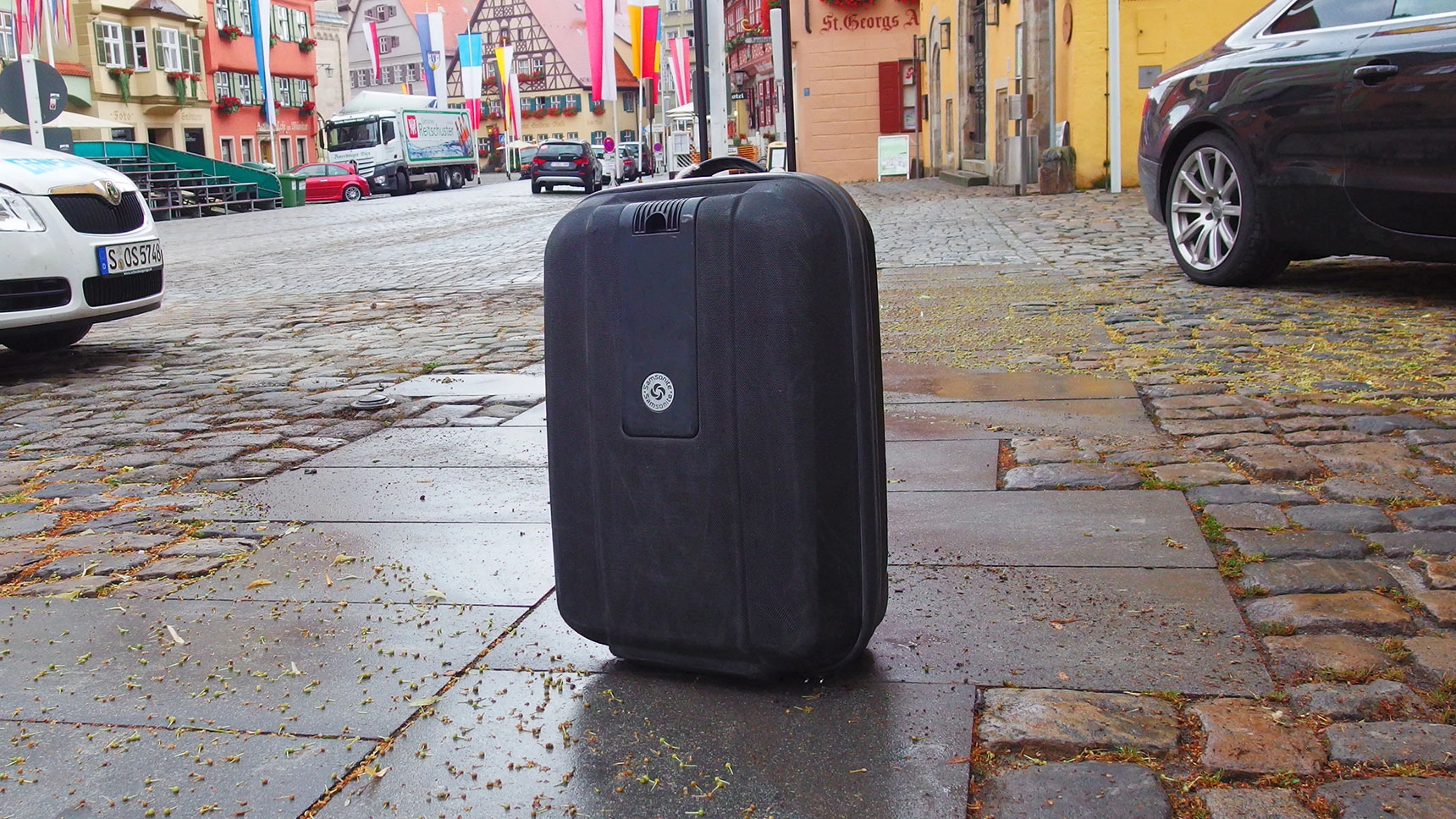 Ein Rollkoffer steht auf einen Gehweg, der teils gepflastert, teils mit Platten belegt ist.