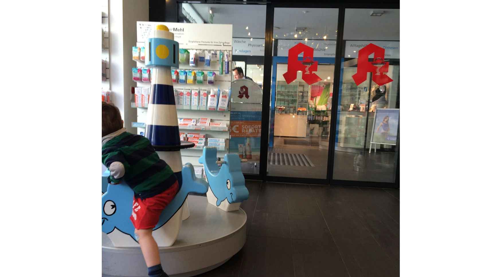 Eingang zu einer Apotheke mit breiter, automatischer Tür. Neben der Tür steht ein Kinderkarussell.