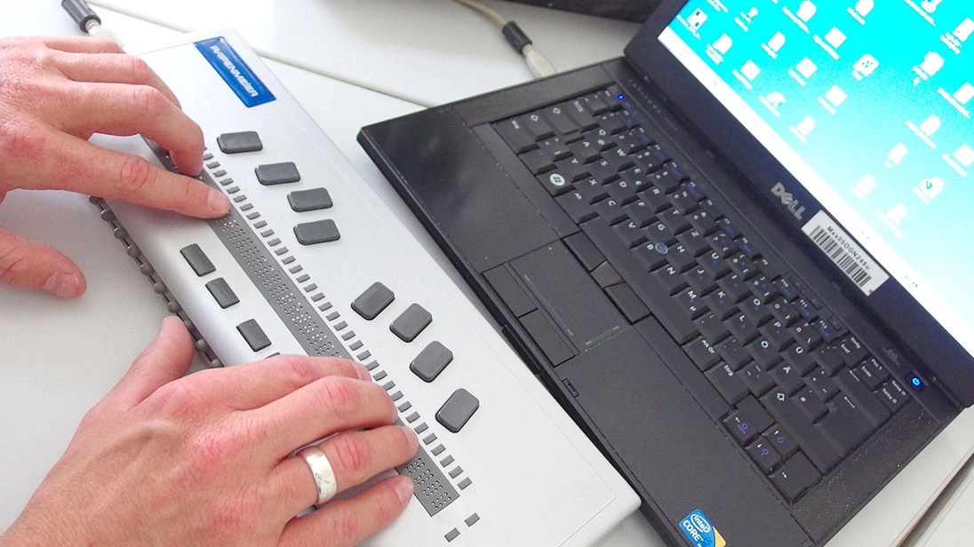 Notebook mit angeschlossenem Brailledisplay. Die Hände des Benutzers liegen auf der Braillezeile.